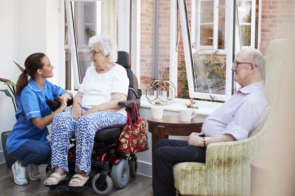 Male And Female Residents Sitting In Chair And Talking With Nurse In Retirement Home